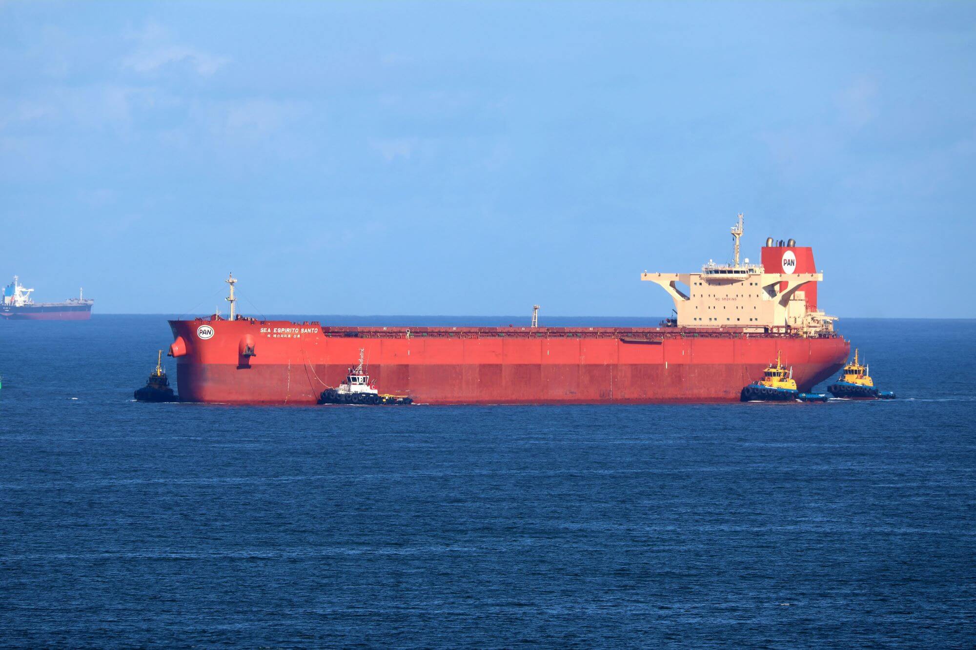 A large red cargo ship being guided by two tugboats on the open sea under a clear blue sky.