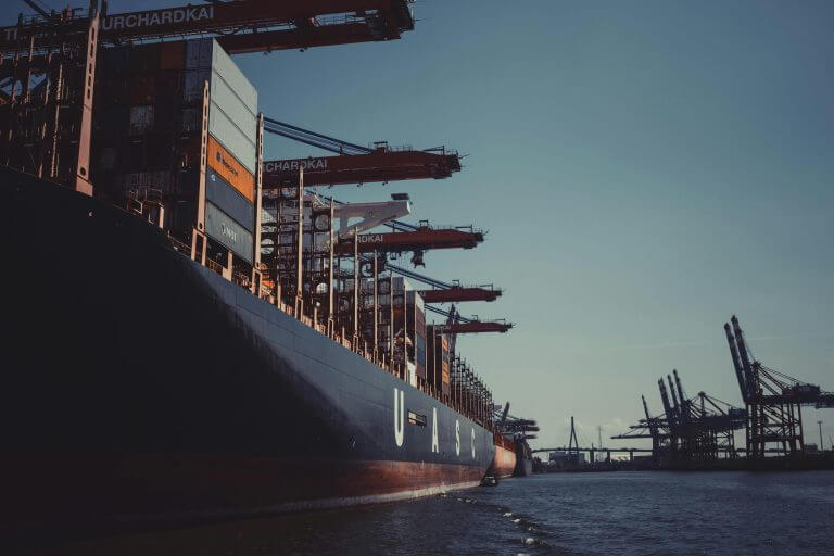 Cargo ship loaded with containers docked at a busy industrial port with cranes ready for unloading during dusk.