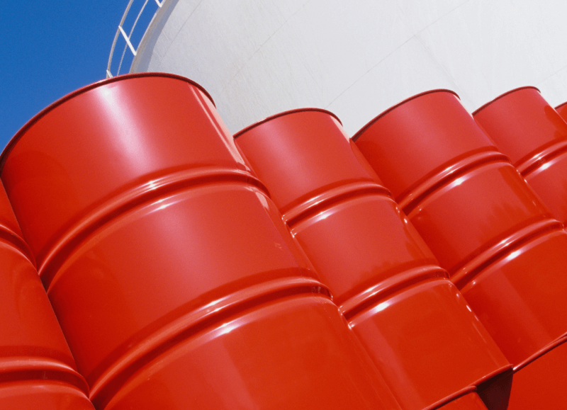 Stack of vibrant red oil barrels labeled with "marine bunker" against a blue sky with a part of a storage tank visible in the background.