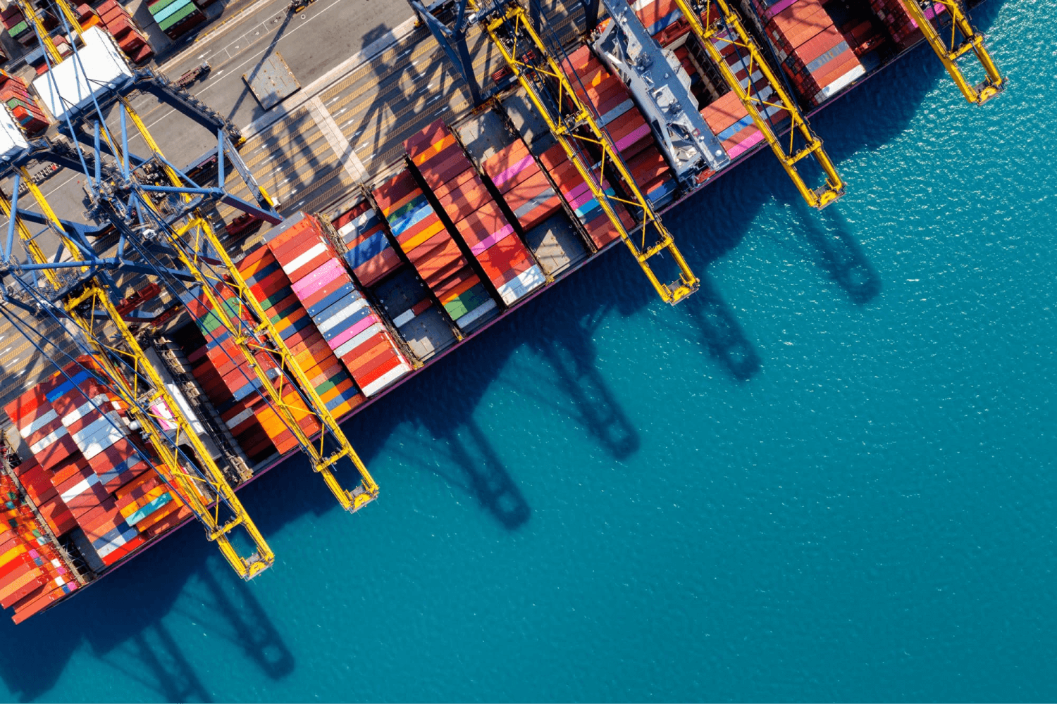 Aerial view of a cargo ship at port being loaded or unloaded by towering yellow gantry cranes, with neatly stacked multicolored shipping containers creating a vibrant pattern on the dock as it prepares to