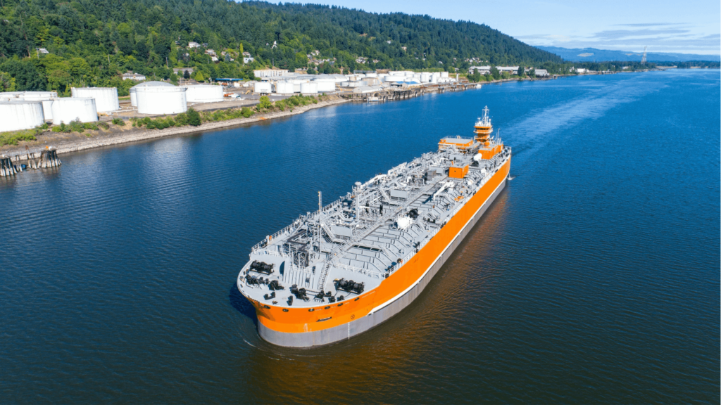 A large orange cargo ship using alternative fuel navigates calm waters near industrial storage facilities under a clear blue sky.