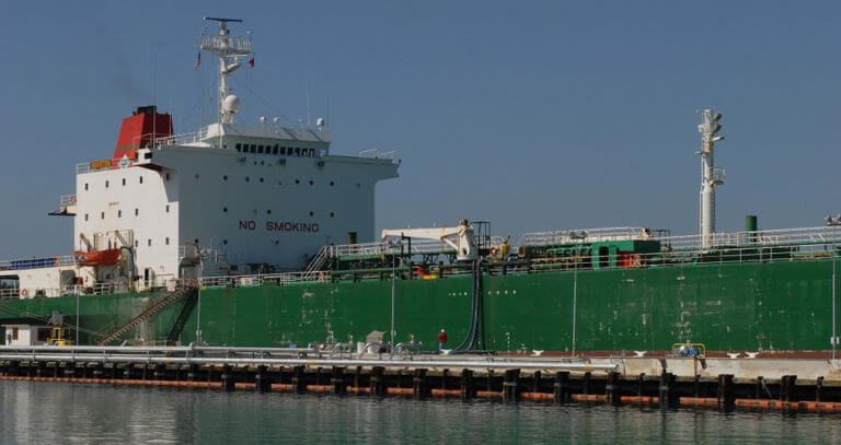 A large, green cargo ship docked at a pier under a clear blue sky, with a prominent "no smoking" sign displayed on its side to ensure safety in the vicinity of the vessel.