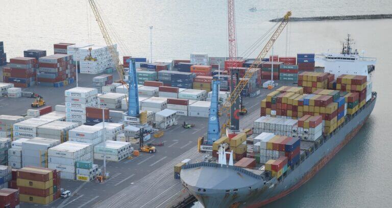 Aerial view of a busy cargo port showing a large freight ship being loaded or unloaded with colorful shipping containers, with cranes and infrastructure visible.