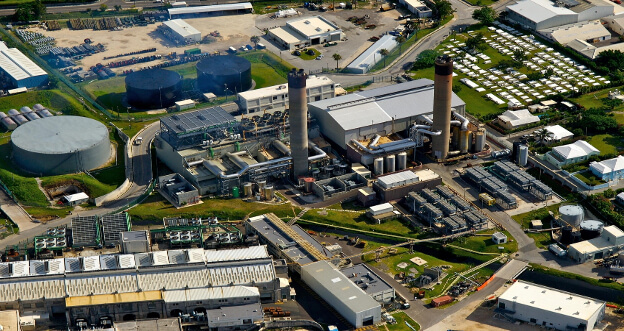 Aerial view of an industrial complex with storage tanks and tall chimneys amidst smaller buildings.