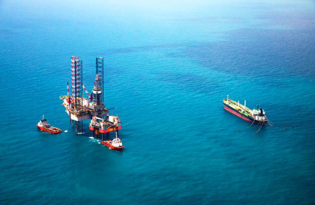 An offshore oil drilling rig flanked by support vessels and a nearby tanker ship in a calm blue sea.