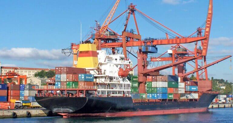 A cargo ship being loaded or unloaded at a bustling commercial shipping dock with large red gantry cranes.