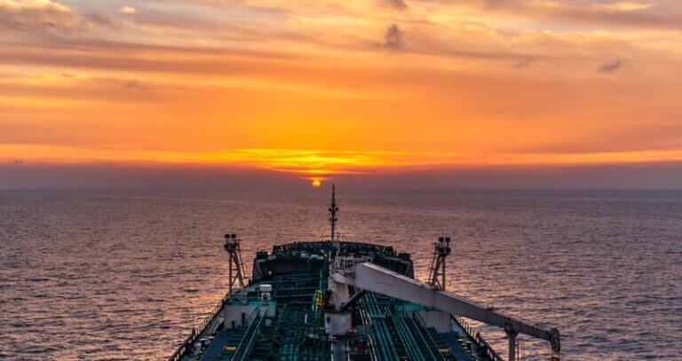 A tanker sailing at sunset, with the vessel's silhouette set against a vibrant orange sky reflecting over a calm sea.